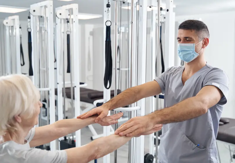 Work Injury Rehab: Masked therapist assists patient. Fce physical therapy for pain relief and mobility recovery at our rehabilitation clinic.